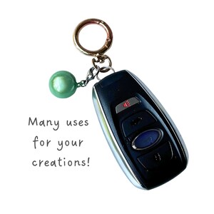 May include: A black car key fob attached to a gold-toned metal keyring with a single round, glossy mint green bead charm. The text "Many uses for your creations!" is printed in a simple dark grey font to the left of the key fob and charm.