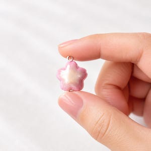 May include: A small, five-petaled flower charm with a glossy finish. The petals transition from a soft pink at the edges to a pearlescent, creamy white center. A small silver-toned metal jump ring is attached to the top of the flower for hanging.