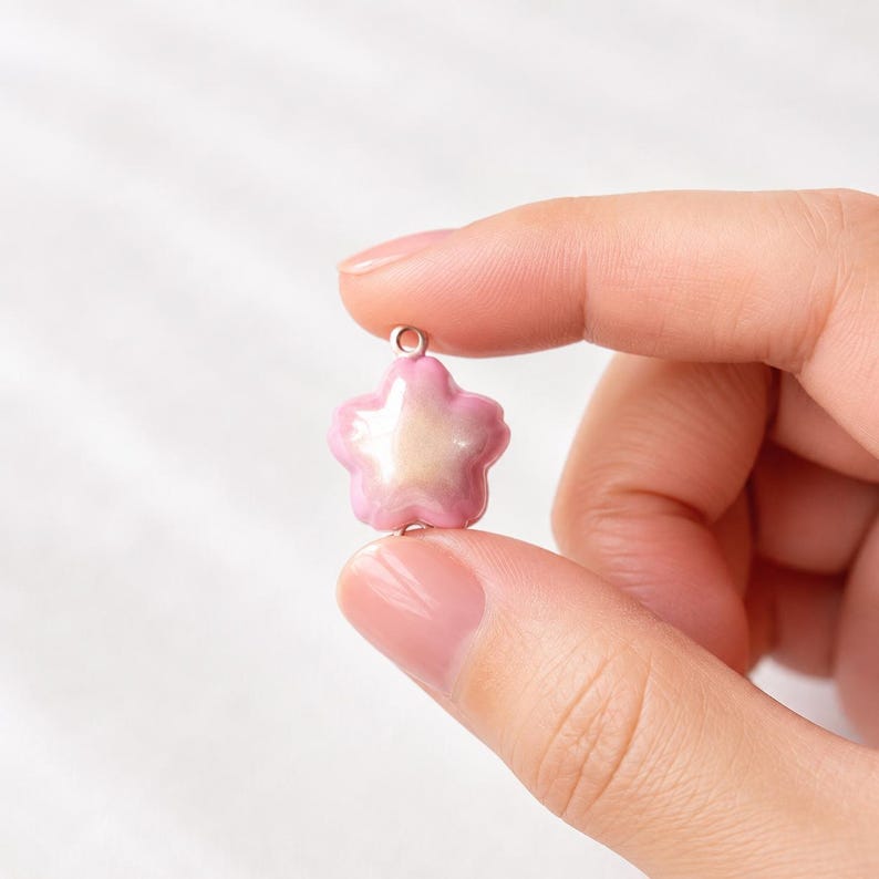 May include: A small, five-petaled flower charm with a glossy finish. The petals transition from a soft pink at the edges to a pearlescent, creamy white center. A small silver-toned metal jump ring is attached to the top of the flower for hanging.