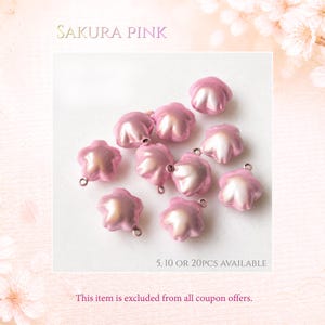 May include: A collection of small, star-shaped charms in a pearlescent sakura pink finish. Each charm features a smooth, rounded surface and a small metal loop at the top for attachment. The text SAKURA PINK, 5, 10 OR 20PCS AVAILABLE is visible.