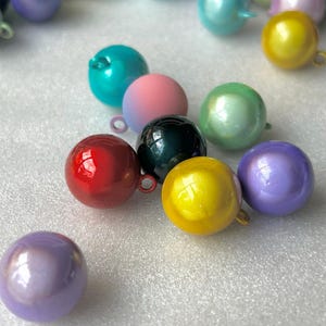 May include: A collection of spherical charms in various colours including red, yellow, teal, pink, black, purple, and green. Each round bead features a small metal loop at the top for attachment, with a smooth, glossy finish reflecting light.