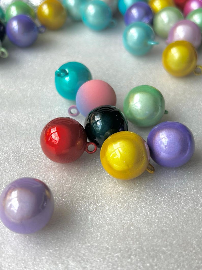 May include: A collection of spherical charms in various colours including red, yellow, teal, pink, black, purple, and green. Each round bead features a small metal loop at the top for attachment, with a smooth, glossy finish reflecting light.