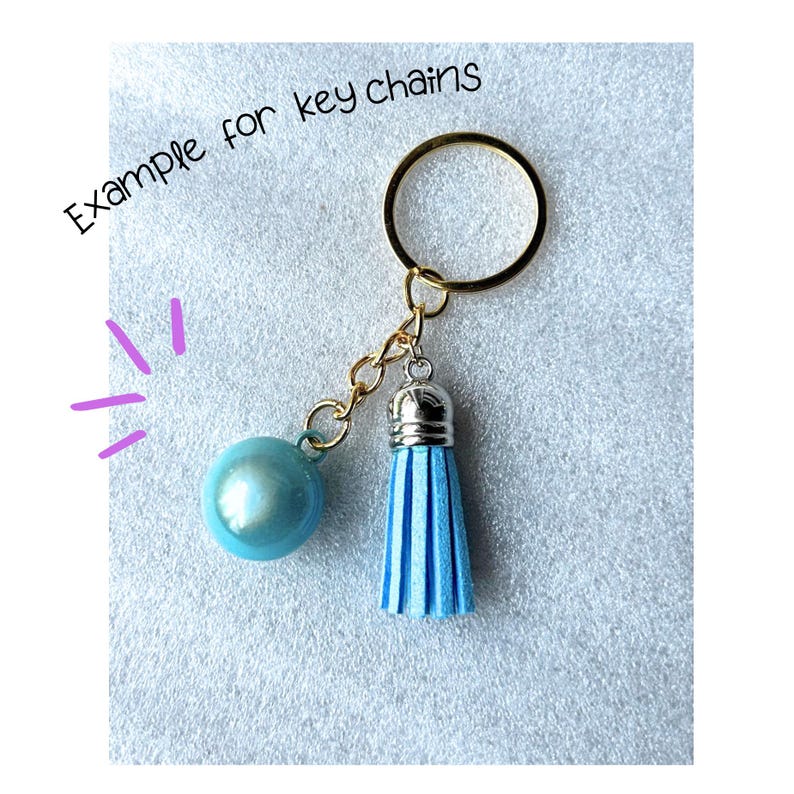 May include: A gold-toned metal keyring featuring a light blue spherical bead and a light blue tassel with a silver-toned cap. The text 'Example for key chains' is printed in black above the object, with purple graphic lines to the left.