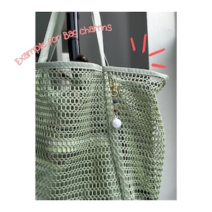 May include: A light green mesh tote bag featuring a gold-toned lobster clasp bag charm with small multicoloured beads and a single white spherical bead. The text "Example for Bag charms" is written in pink script across the upper left corner.