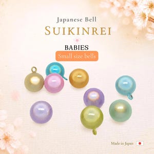 May include: A collection of small, spherical Japanese Suikinrei bells in various pastel colors including light blue, pink, purple, gold, and green. Each bell features a small metal loop at the top for attachment, with text reading Japanese Bell SUIKINREI.