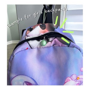 May include: A purple and blue gradient backpack featuring a cartoon character print and the text "Example for girls backpacks" in white. The bag has a black zip closure with a round, light green charm attached to the metal zip pull.