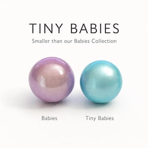 May include: Two smooth, spherical objects with a pearlescent finish. One is light purple, labeled 'Babies', and the other is light blue, labeled 'Tiny Babies'. The text 'TINY BABIES' and 'Smaller than our Babies Collection' appears above them.