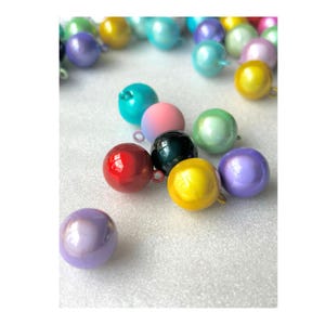 May include: A collection of spherical charms in various colours including red, yellow, purple, teal, pink, and green. Each round bead features a small metal loop at the top for attachment. The surface of these charms has a smooth, glossy finish.