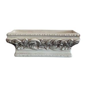 Puede incluir: Una jardinera rectangular decorativa de color blanquecino con adornos florales en relieve. La jardinera tiene una superficie texturizada y un borde de cuentas a lo largo del borde superior. La base es de forma rectangular.