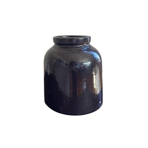 Op de afbeelding: Een donkere, glanzende keramische pot met een afgeronde body en een kleine, cilindrische hals. De pot heeft een glad, reflecterend oppervlak en de kleur is een diep, rijk zwart. De pot is een decoratief item.