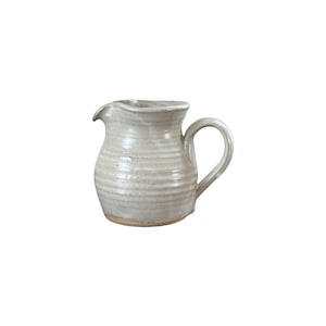 May include: A small, off-white ceramic pitcher with a handle and a spout. The pitcher has a rounded body with horizontal ridges and a slightly textured surface. The handle is curved, and the spout is gently curved.