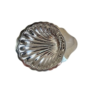 Könnte beinhalten: Eine polierte, silberfarbene Metallschale in Form einer Jakobsmuschel. Die Schale weist eine Reihe tiefer, strahlenförmig verlaufender Rillen auf, die von der Basis bis zum gewellten Rand reichen und eine reflektierende Oberfläche mit Griff bilden.