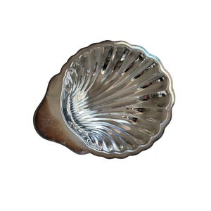 Può includere: Un piatto in metallo color argento lucido a forma di conchiglia di capasanta. Il piatto presenta una serie di creste e scanalature radianti che si estendono dalla base al bordo smerlato, creando una superficie riflettente con finitura metallica.