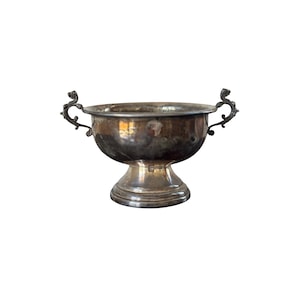 May include: A silver-toned, antique bowl with a pedestal base and two ornate handles. The bowl has a wide, shallow basin and shows signs of age and wear. The handles are intricately designed with a scrolled pattern. The bowl is likely made of metal.