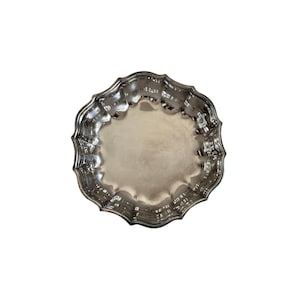 Könnte beinhalten: Ein rundes, silberfarbenes Metalltablett mit einer glatten, reflektierenden Mitte und einem dekorativen, gewellten Rand mit mehreren geschwungenen Vertiefungen. Die Oberfläche hat ein poliertes Finish, das das Licht einfängt.