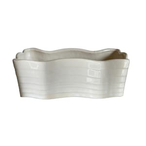Puede incluir: Una jardinera rectangular de cerámica blanquecina con borde superior ondulado y detalles de estrías horizontales. La jardinera tiene un acabado brillante. El diseño es sencillo y elegante.