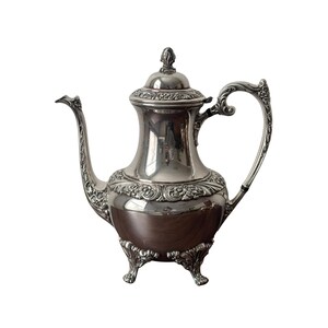 May include: A silver-plated coffee pot with ornate detailing. The pot features a curved spout, a decorative handle, and a lid with a finial. The lower portion of the pot has a dark brown finish, and it stands on three decorative feet.