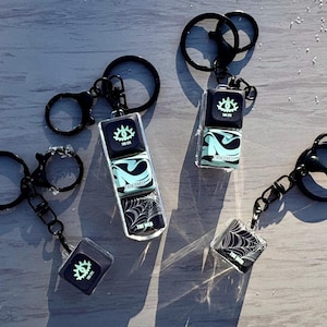 May include: Black keychains with a variety of designs. One keychain has three stacked, clear cubes with a glowing eye, a swirling design, and a spiderweb. Other keychains have a single cube with the same eye design or spiderweb design. The text "THE WEB" is visible.