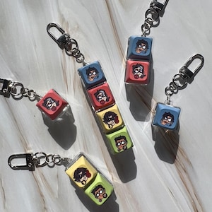 May include: Keychains featuring colorful, square charms with cartoon character illustrations. The charms are in various colors, including blue, red, yellow, and green, and are attached to silver-tone key rings. The keychains are displayed on a light-colored surface.