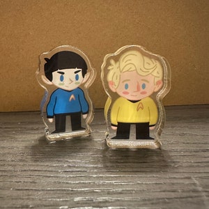 May include: Two small, cartoon-style figurines of characters from Star Trek. The figure on the left is wearing a blue uniform with a gold emblem. The figure on the right is wearing a yellow uniform with a gold emblem. Both figures are standing on a wooden surface.