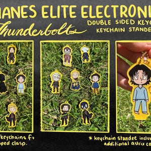 May include: The image showcases double-sided keychains and keychain standees featuring various character designs. The keychains have a yellow border and a star-shaped clasp. The text "SHANES ELITE ELECTRONIC... Thunderbolts" is visible at the top.