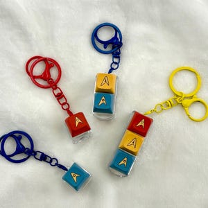 May include: Keyrings with keycaps in red, blue, and yellow, each displaying the Star Trek insignia. The keycaps are clear with the letter "A" and a star symbol. The keyrings have metal clasps in matching colours.