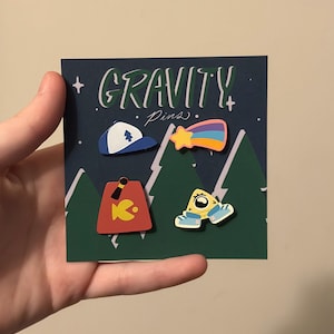 Könnte beinhalten: Ein Set mit vier Gravity Falls Emaille-Pins. Die Pins zeigen einen blauen Hut mit einem weißen Dreieck, einen gelben Sternschnuppen mit einem Regenbogen, einen roten Fez mit einem gelben Fisch und ein gelbes Wesen mit blauen Schuhen.