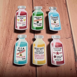 Op de afbeelding: Zes stickers in de vorm van serumflessen. Elke fles heeft een ander kleurenschema en tekst, waaronder "Super Soldier Serum", "C.I.A. Top Secret" en "Project Sentry". De flessen zijn gerangschikt op een houten oppervlak.