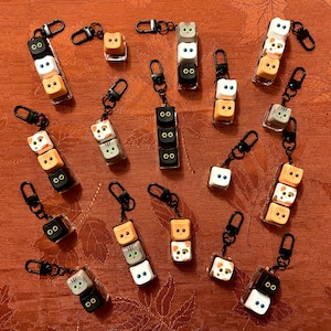 May include: A collection of cat-shaped keychains with black clips. Each keychain features stacked, square blocks with cat faces in various colors, including white, orange, gray, and black. The keychains are displayed on a reddish-orange surface.