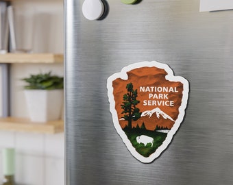 May include: A magnet featuring the National Park Service logo. The design includes a mountain range, a tree, and a bison, all in a vintage style. The text "NATIONAL PARK SERVICE" is prominently displayed.