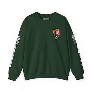 Op de afbeelding: Een donkergroene sweatshirt met het logo van de National Park Service op de borst. De mouwen hebben witte strepen en de woorden "National Parks" zijn verticaal gedrukt. De sweatshirt heeft een ronde hals.