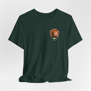 May include: Dark green t-shirt with the National Park Service logo on the left chest. The logo is an arrowhead design with a tree, mountain, and bison illustration. The text "National Park Service" is above the illustration.