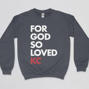 Pode incluir: Um moletom cinza escuro com as palavras "FOR GOD SO LOVED KC" impressas em branco e vermelho. O moletom tem mangas compridas e gola redonda. O texto está alinhado verticalmente na frente.