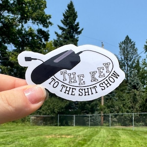 May include: A white sticker with a black computer mouse graphic and the text "THE KEY TO THE SHIT SHOW" in a black, oval design. The sticker is held up against a background of green grass and trees.