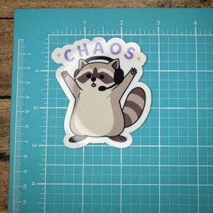 May include: A white sticker featuring a cartoon raccoon wearing a headset and raising its arms. The word "CHAOS" is written above the raccoon in purple letters with yellow stars. The sticker is on a blue grid.