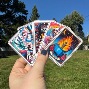 May include: A hand holds a fan of five tarot card stickers. Each card features colorful illustrations with themes of skulls, flowers, and celestial elements. The text "NOT ENOUGH" and "THE SHIFT" are visible on two of the cards.