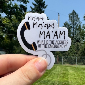 May include: A white sticker with a black telephone receiver graphic and the text "Ma'am Ma'am MA'AM WHAT IS THE ADDRESS OF THE EMERGENCY?" The receiver has a yellow stripe. The sticker is held up against a background of green trees and blue sky.