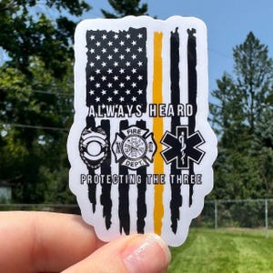 May include: A sticker featuring a distressed American flag design with a yellow stripe. The sticker includes the text "ALWAYS HEARD PROTECTING THE THREE" and symbols for law enforcement, fire department, and emergency medical services.