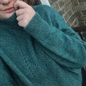 Może przedstawiać: Sweter z dzianiny w kolorze turkusowym z delikatnym wzorem ażurowym. Sweter ma wysoki kołnierz i długie rękawy.