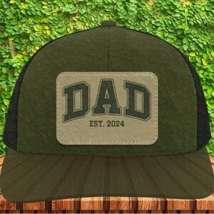 May include: A green and black trucker hat with a brown patch that says "DAD EST. 2024".