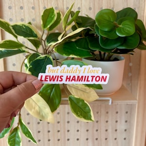 May include: A sticker with the text "but daddy I love LEWIS HAMILTON" is held in front of two potted plants. The sticker has a white background with red and yellow text. The plants have green and yellow leaves and are in white pots.