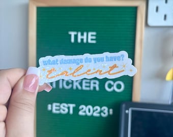 May include: A sticker with the text "what damage do you have? talent" in blue and orange, with the words "STICKER CO" and "EST 2023" below. The sticker is held in front of a green felt board.