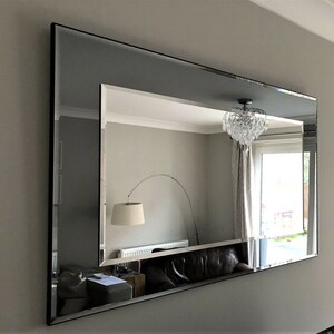 May include: A large, rectangular mirror with a silver frame. The mirror is hanging on a wall and reflects a room with a chandelier, curtains, and a sofa.