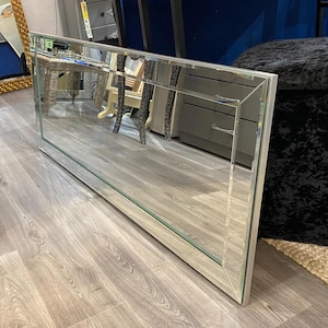 May include: A large rectangular mirror with a silver frame and beveled edges. The mirror reflects the room, including furniture and a wooden floor. The frame has a geometric design, adding a modern touch to the decor.