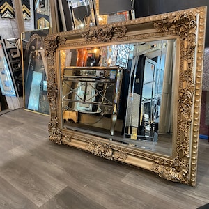 May include: An elaborate gold-framed mirror with detailed floral and scroll patterns. The mirror reflects a room with other mirrors and furniture. The frame has a distressed, antique look, enhancing its vintage appeal. A decorative home decor item.