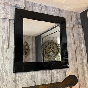 May include: Square black framed mirror. The mirror is mounted on a wall with a distressed wood panel design. The reflection shows a room with a large clock and other decorative elements.