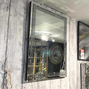 May include: A rectangular mirror with a black frame and a decorative border of sparkling crystals. The mirror is hanging on a grey wooden wall, reflecting the room's interior. The mirror's design adds a touch of glamour to the space.