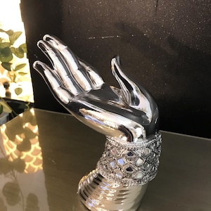 May include: A silver decorative hand sculpture with a polished finish. The wrist area has a band with small, mirrored tiles. The hand is in an open gesture, suitable as a display piece or decorative accent. The sculpture is likely made of metal.