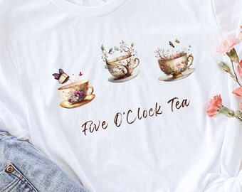 Puede incluir: Camiseta blanca con un diseño "Five O'Clock Tea" que presenta tres tazas de té con detalles florales y de mariposas. La camiseta se combina con vaqueros de mezclilla de lavado claro y un sombrero de paja. Flores rosas dispuestas a un lado.