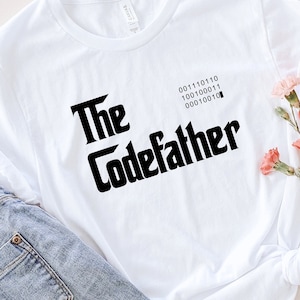 Op de afbeelding: Wit T-shirt met de tekst "The Codefather" in een zwarte, gestileerde lettertype. Het shirt heeft ook binaire code en wordt gecombineerd met een lichtblauwe jeans en een strohoed. Roze bloemen voegen een vleugje kleur toe.